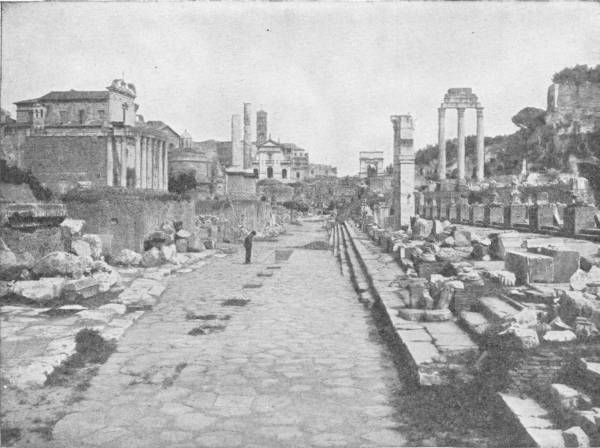 The Forum at Rome as It Is To-Day — A paved Roman road runs straight between broken columns and fallen blocks of stone, with three standing columns on the right, a pillared temple front on the left and a single figure far down the way.