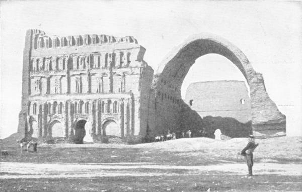 The Great Roman Arch at Ctesiphon Near Bagdad — A ruined brick hall with rows of blind arches along its front stands beside one enormous open arch, with small figures at its foot and a man standing in the foreground.