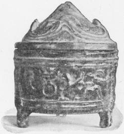A Chinese Covered Jar of Green-Glazed Earthenware. Han Dynasty (contemporary with the late Roman republic and early Empire) (In the Victoria and Albert Museum) — A round jar standing on three short feet, with a band of figures in low relief around its body and a peaked lid shaped like a cluster of hills.