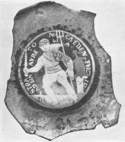 A contemporary picture of a gladiator: a round medallion in a broken piece of glass shows a man in a short tunic holding a weapon and a staff, with Latin letters around the rim.