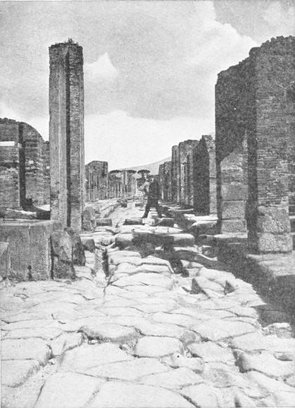 Pompeii. “Note the ruts in roadway worn by chariot wheels.” — A paved street with deep ruts running between the brick walls of roofless houses, with raised stepping-stones across the road and a man standing among the ruins.
