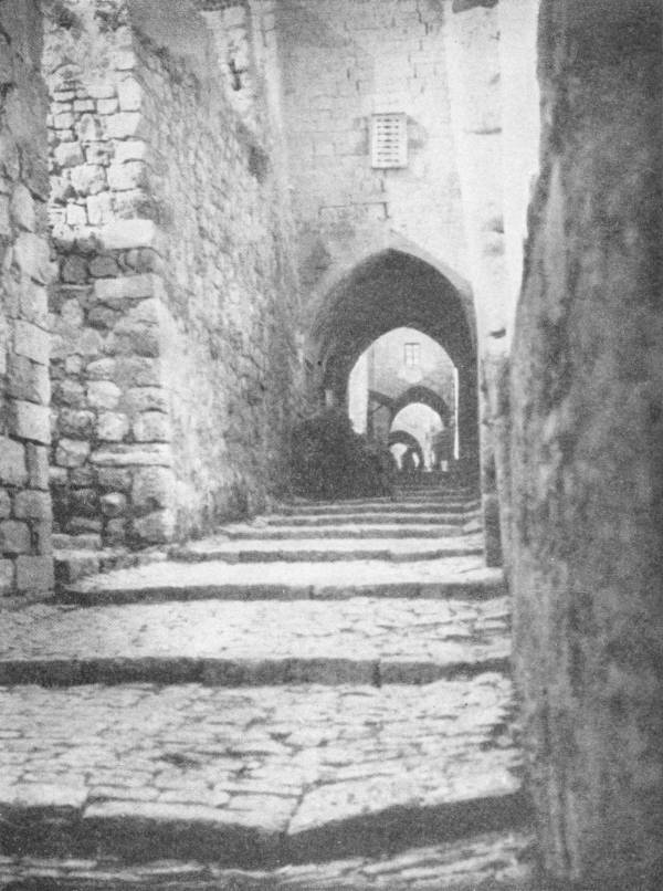 A Street in Jerusalem. Along such a thoroughfare Christ carried his cross to the place of execution. Photo: Fannaway — A narrow stepped street of worn paving climbs between high stone walls and passes under a series of pointed arches.