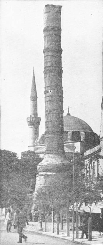Constantine’s Pillar, Constantinople. Photo: Sebah & Foaillier — A tall, battered column built up of stone drums stands on a broad base in a street, with a mosque’s dome and minaret behind it.