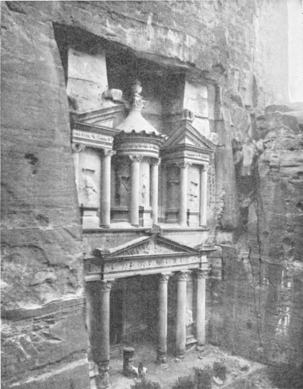 The Rock Hewn Temple at Petra. Photo: Underwood & Underwood — A temple front cut straight into the face of a cliff, in two storeys of columns, with a round pavilion under a pointed roof at the top and a small figure at its foot.