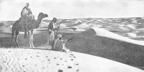 Looking Across the Sea of Sand. Photo: Lehnert & Landrock — On the top of a dune a man on a camel and a man with a staff look on while a third kneels and points out over rolling waves of sand that reach to the horizon.