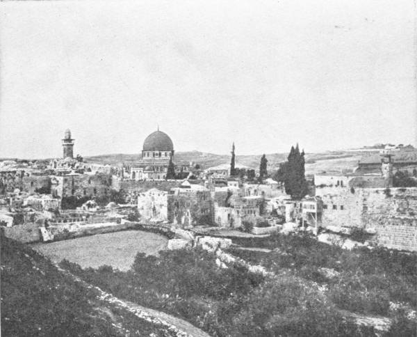 Jerusalem, Showing the Mosque of Omar. Photo: Lehnert & Landrock — A photograph across open ground and flat roofs to the walled city, where the great dome of the mosque rises above the houses, with a square minaret to its left and dark cypresses to its right.