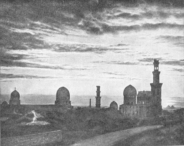 View of Cairo Mosques. Photo: Lehnert & Landrock — The domes and tall minarets of a row of mosques stand in silhouette against a streaked evening sky, with a road curving away in the foreground.