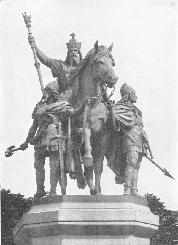 Statue of Charlemagne in Front of Notre Dame, Paris. The figure is entirely imaginary and romantic. There is no contemporary portrait of Charlemagne. Photo: Rischgitz — A bronze statue on a tall stone plinth: the crowned, bearded emperor rides a walking horse holding a tall sceptre, led by two helmeted men in mail, one carrying an axe and the other a spear.