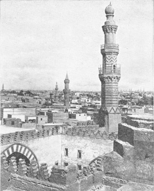 View of Cairo. Photo: Lehnert & Landrock — A view over the flat roofs of the city from a ruined wall with a striped arch, a tall banded minaret with balconies in front and more minarets behind.