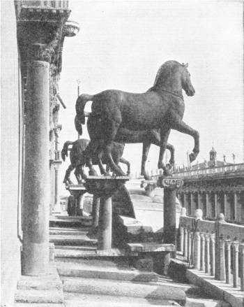 The Horses of S. Mark, Venice. Originally on the arch of Trajan at Constantinople, the Doge Dandalo V took them after the Fourth Crusade, to Venice, whence Napoleon I removed them to Paris, but in 1815 they were returned to Venice. During the Great War of 1914-18 they were hidden away for fear of air raids. Photo: D. McLeish — Two of the four bronze horses, each with a raised foreleg, stand on stone pillars on the gallery of the church front, with columns and a balustrade around them.