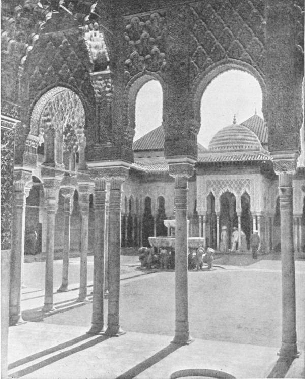 A Courtyard in the Alhambra. Photo: Lehnert & Landrock — A view through carved arches on slender paired columns into a paved courtyard, with a fountain on the backs of stone lions at its centre and a domed pavilion beyond.