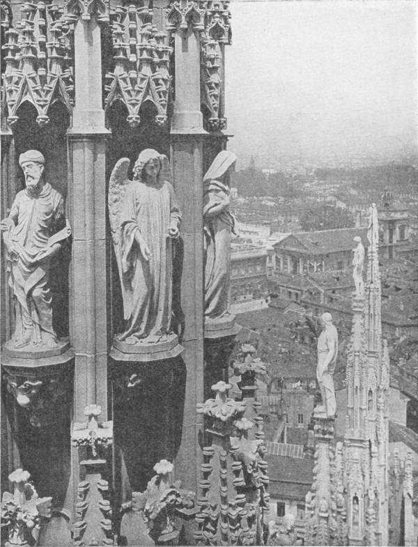 Milan Cathedral. View showing the exquisite carvings characteristic of the 98 spires of the edifice — Carved stone saints and an angel stand in niches under pointed canopies high on the cathedral roof, with pinnacles and statues rising beside them and the city spread out below.