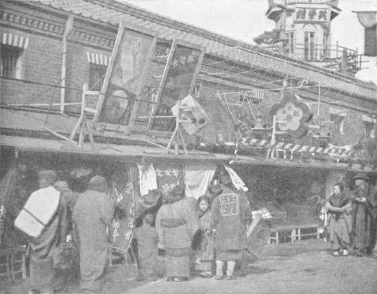 A Street in Tokio — People in kimonos gather at open shopfronts hung with cloth banners, beneath large painted signboards set up along the roof.