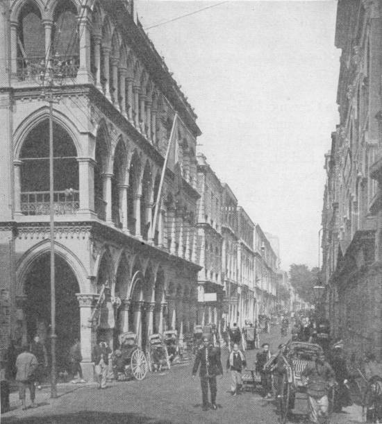 Street in Hong Kong. Photo: Underwood & Underwood — A street of tall arcaded buildings with balconies, where men in European and Chinese dress walk among rickshaws.