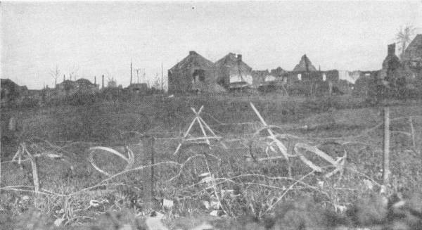 The Devastation of Modern War. Wire entanglements in the foreground. Photo: Photopress — Barbed wire strung on stakes and trestles stretches across a field in front of a row of shattered houses.