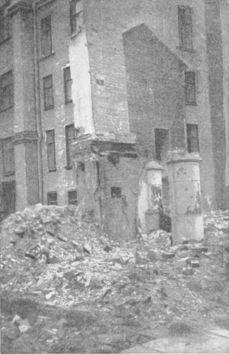 A View in Petersburg Under Bolshevik Rule. A wooden house has been demolished for firewood. By courtesy of Messrs. Hodder & Stoughton — A heap of rubble fills the gap where a house stood, against the bare side wall of a tall block, with two short round pillars at its edge.