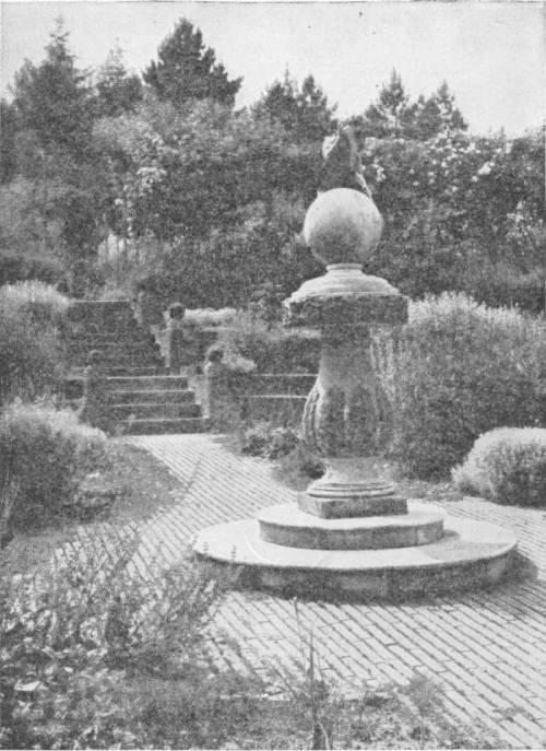 A Peaceful Garden in England. Given wisdom, all mankind might live in such gardens — A stone ball on a carved pedestal stands on a round step in a brick-paved garden, with flowering shrubs, stone steps and tall trees behind.
